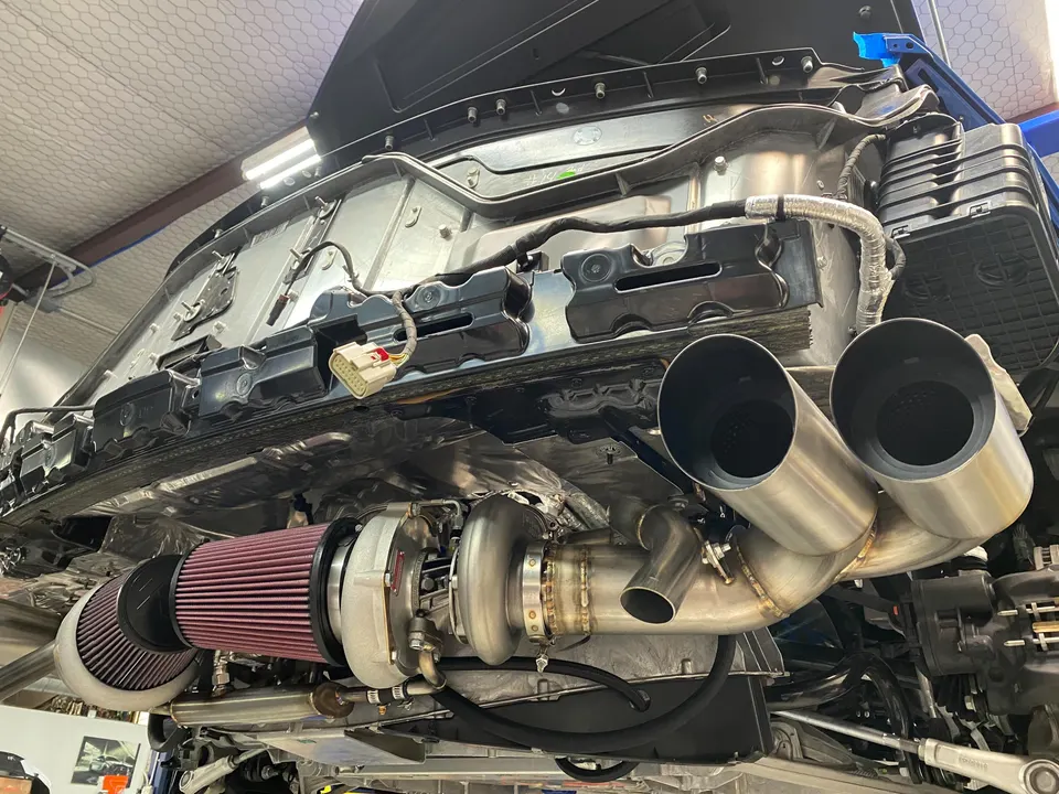 Underside view of a performance car on a lift, showcasing a large turbocharger, custom exhaust system, and air filters.
