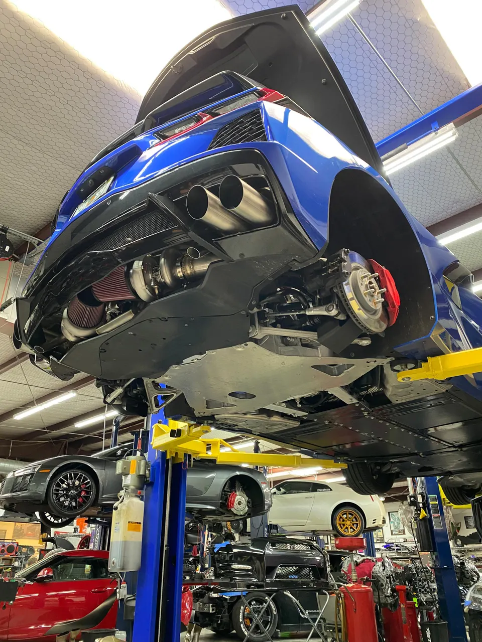 A blue Corvette C8 on a lift in a performance shop, showcasing a custom turbocharger and exhaust system installation.