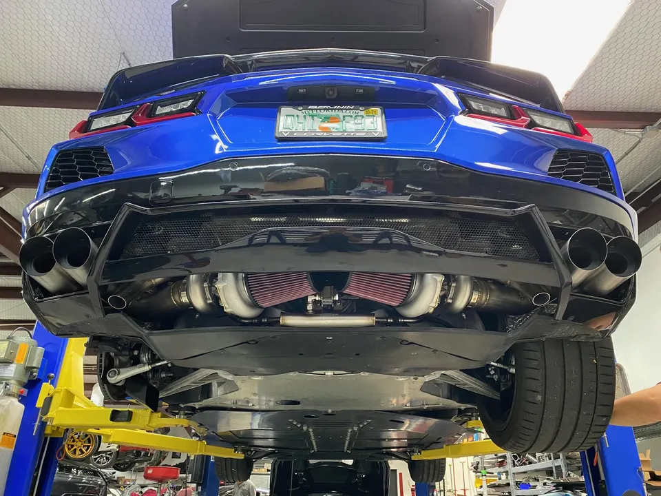 A blue C8 Corvette on a lift in a performance shop, showcasing its twin-turbo setup and custom exhaust system from the rear underside.