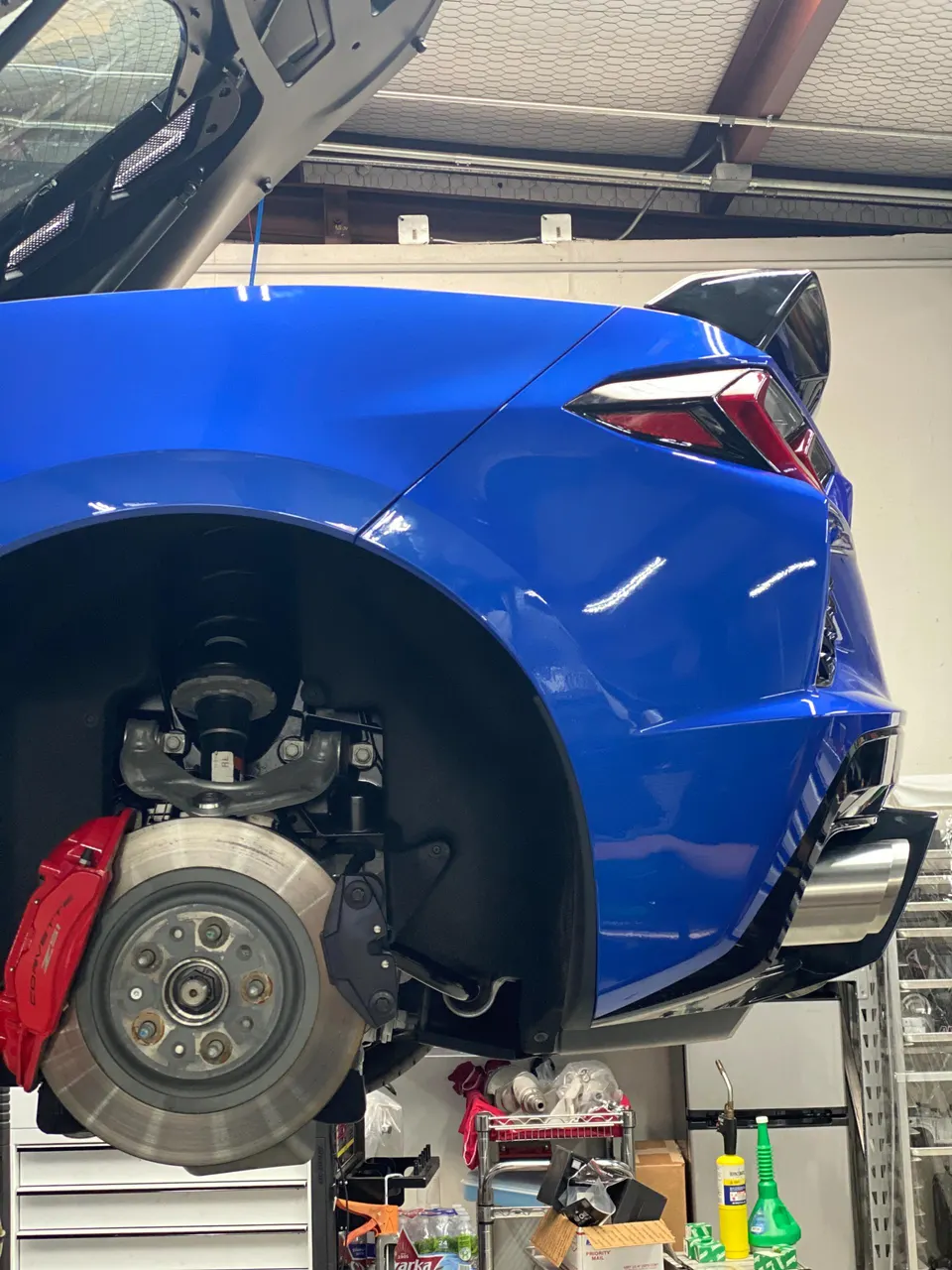 A blue C8 Corvette on a lift in a shop, with the rear wheel removed, showcasing the red brake calipers and suspension.