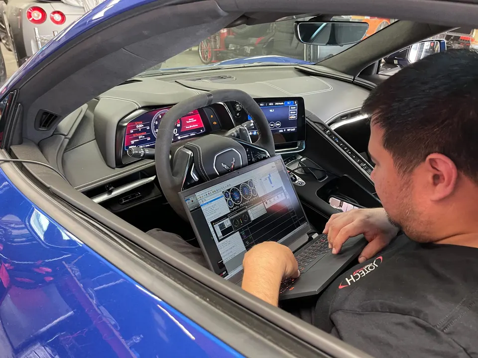 A Jotech technician tunes a blue C8 Corvette's engine using a laptop connected to the car's system.