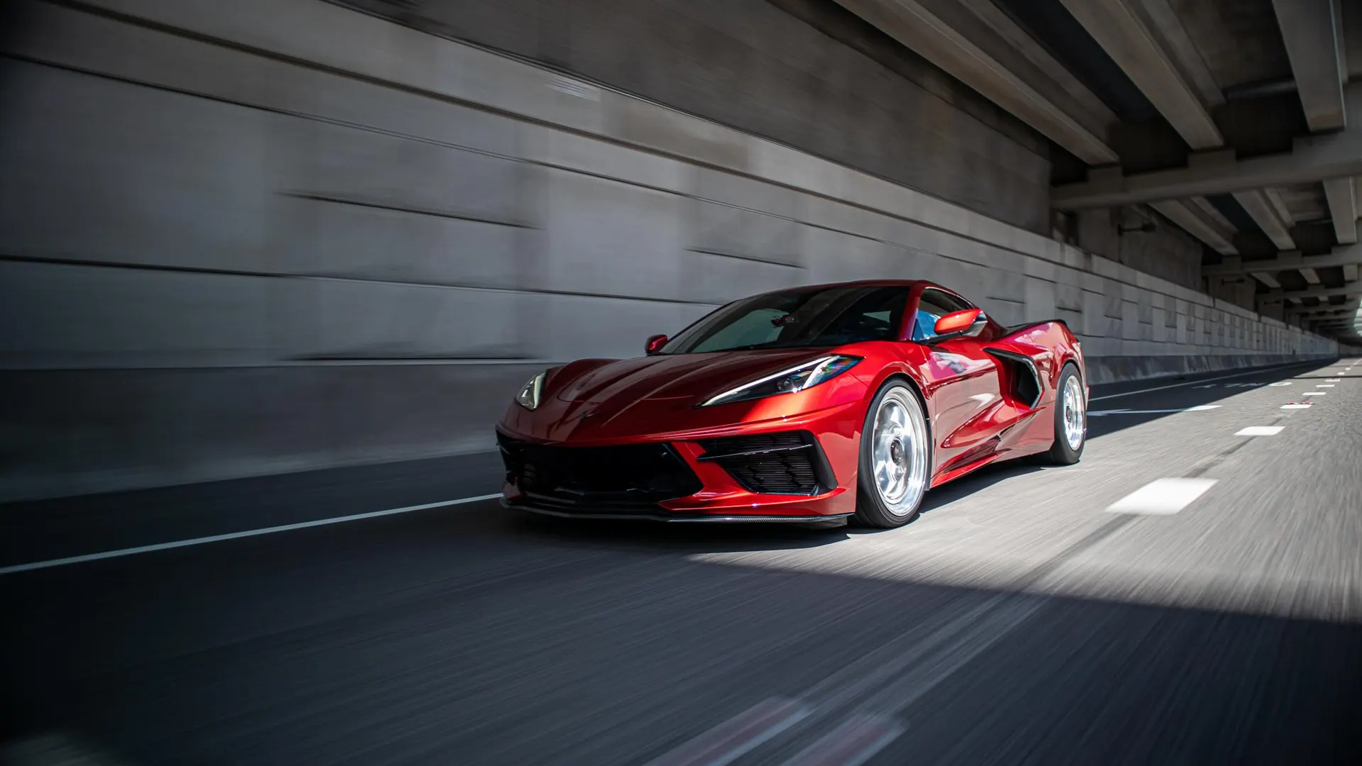 A vibrant red C8 Corvette with custom silver wheels drives dynamically through a concrete tunnel, captured in a professional roller shot.