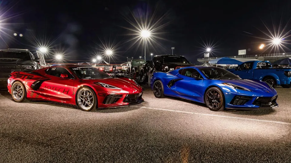 Two Chevrolet C8 Corvettes, one red and one blue, parked under bright lights at a drag strip at night.