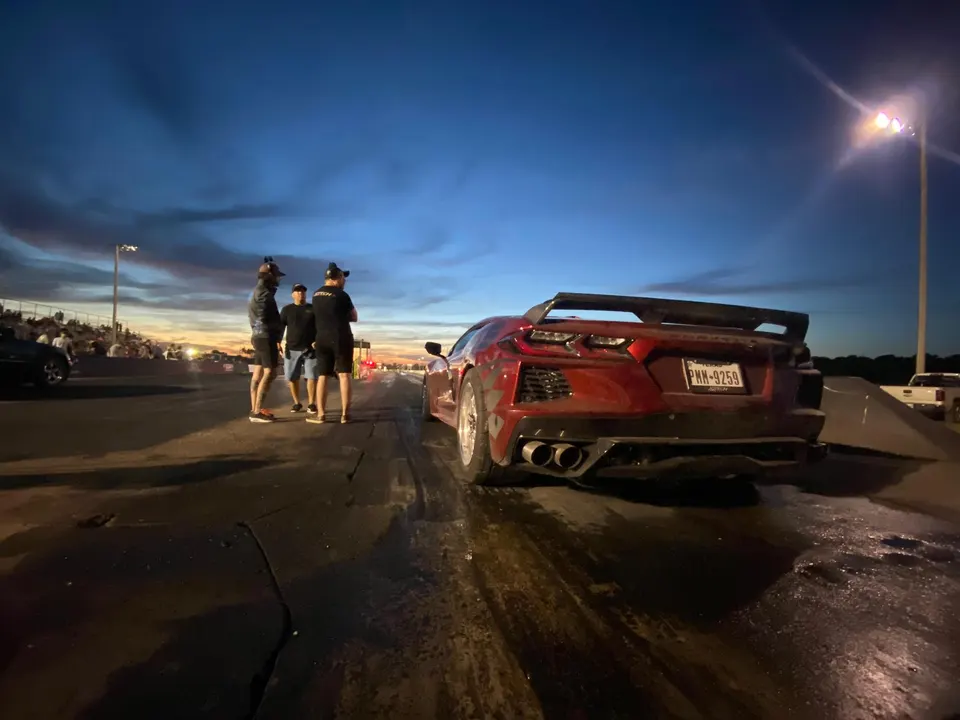 A red C8 Corvette with a large rear wing at a drag strip during sunset, with people standing nearby.