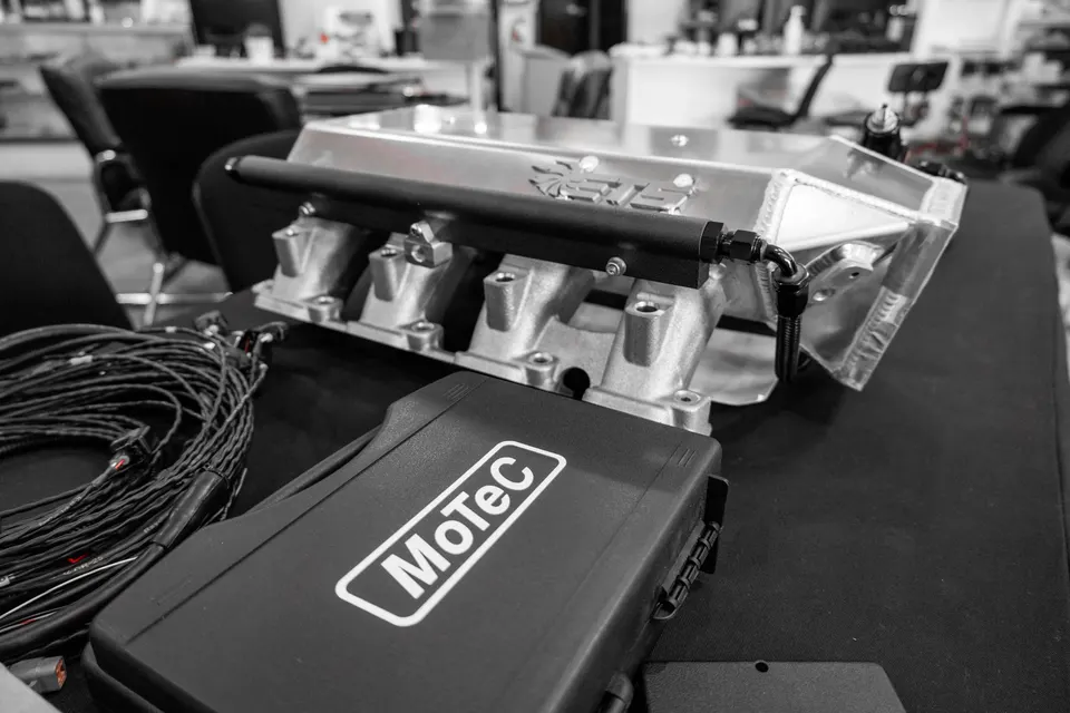 Aftermarket intake manifold, fuel rail, MoTeC ECU, and wiring harness laid out on a table in a performance shop.