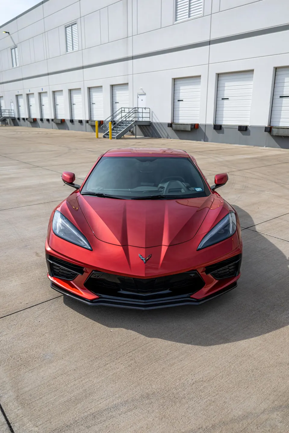 Front view of a red Chevrolet Corvette C8 parked in a warehouse loading dock area on a sunny day.