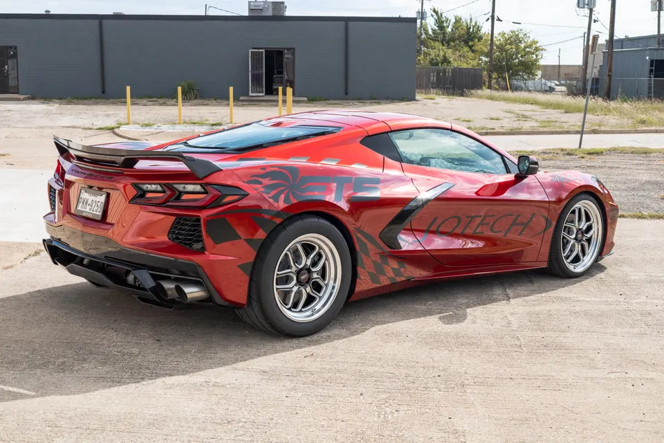 A red Corvette C8 with custom black and red decals, an aftermarket rear spoiler, and drag wheels parked outdoors.