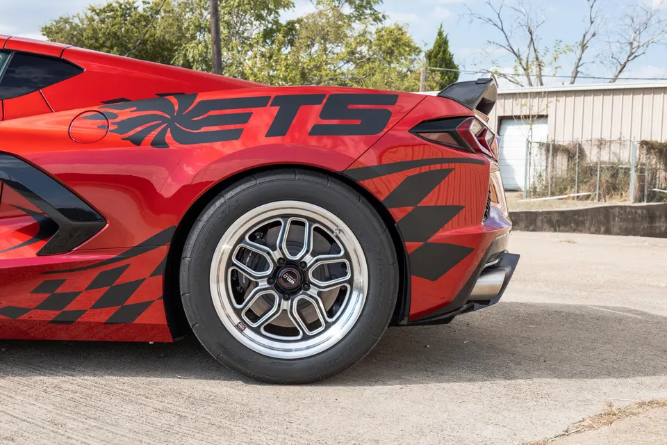 A red Corvette C8 with custom black graphics, aftermarket drag wheels, and performance tires parked outdoors.