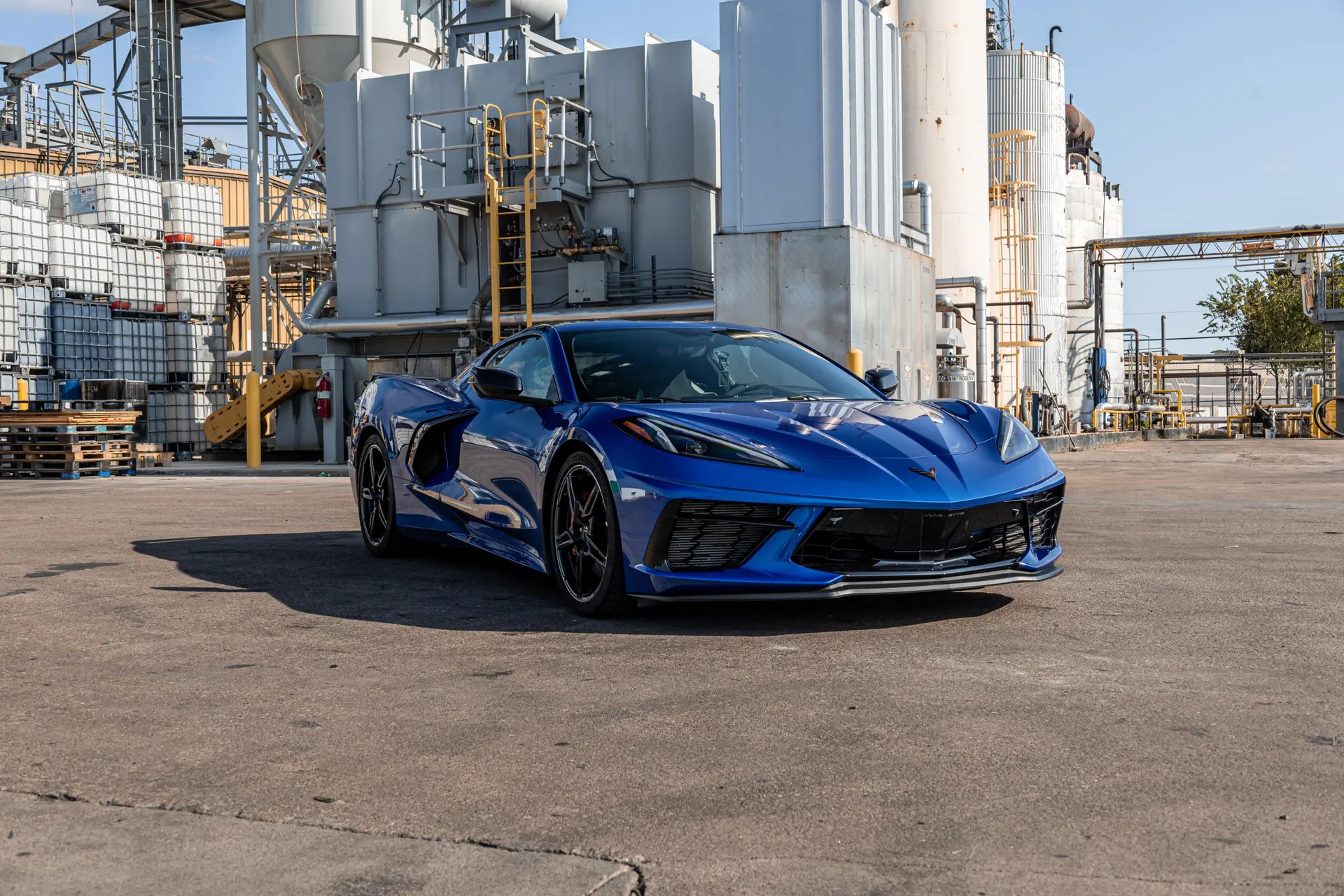 A blue Chevrolet Corvette C8 parked in an industrial area with large tanks and buildings under a clear sky.
