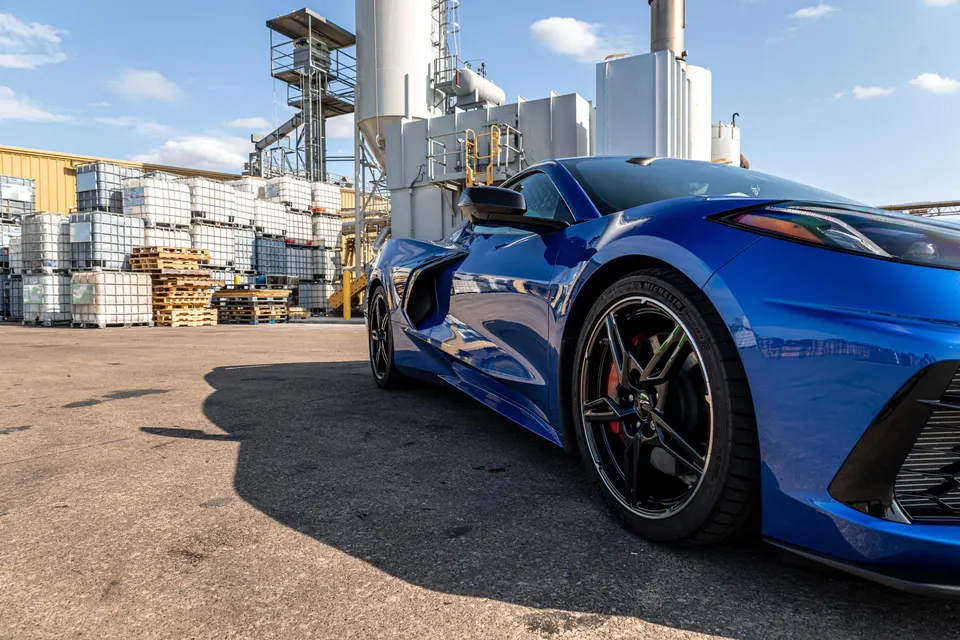 A blue Chevrolet Corvette C8 sports car parked in an industrial yard with stacks of white containers and factory buildings.