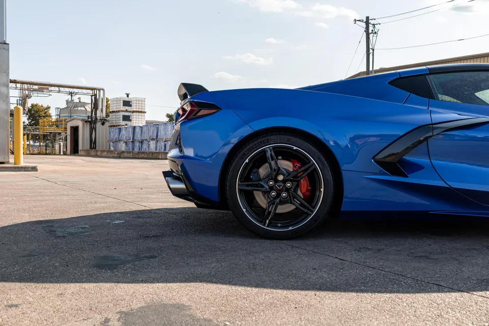 Blue Chevrolet Corvette C8 parked outdoors, showcasing its rear wheel with black rims and red brake calipers, and a rear spoiler.
