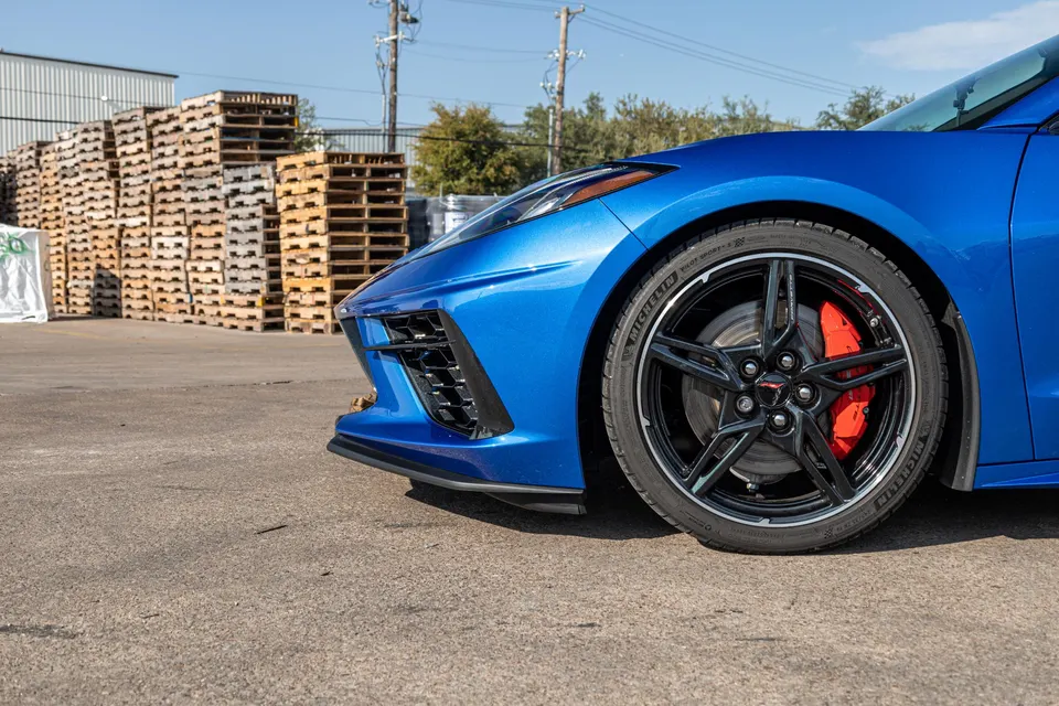 A close-up of the front wheel and fender of a blue C8 Corvette, showcasing black wheels and red brake calipers.