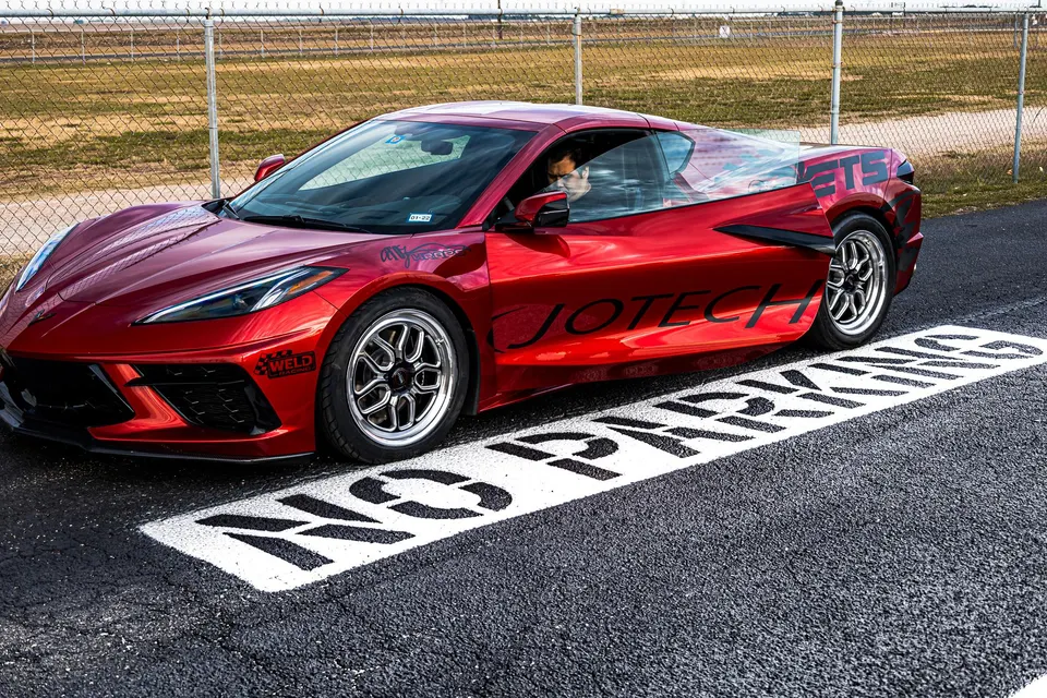 Red C8 Corvette with Weld Racing drag wheels parked on an asphalt track surface next to a chain-link fence.