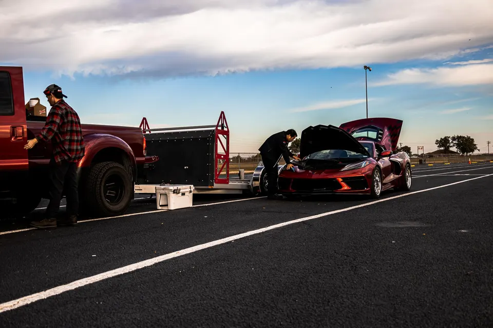 Red Corvette C8 with open hoods being prepped at a track, parked next to a red truck and trailer under a cloudy sky.