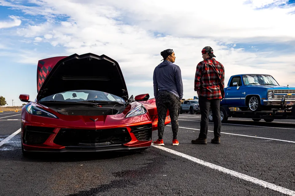 Two men stand next to a red Corvette C8 with its front trunk open at an outdoor car event, with a classic blue Chevrolet truck on a trailer in the background.