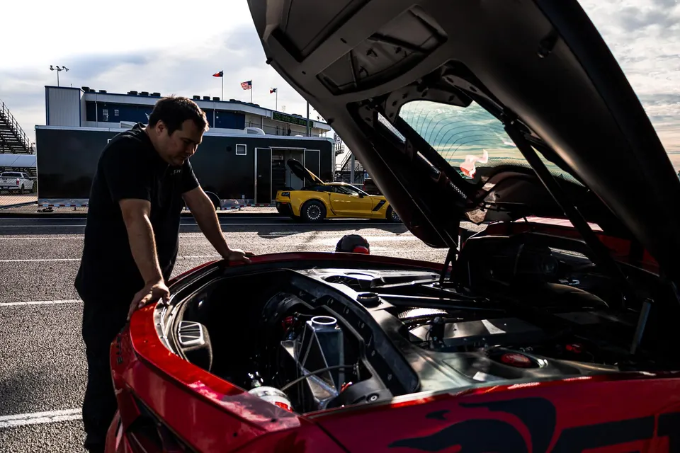 A man inspects the open engine bay of a red Corvette at a drag strip, with a yellow Corvette in the background.
