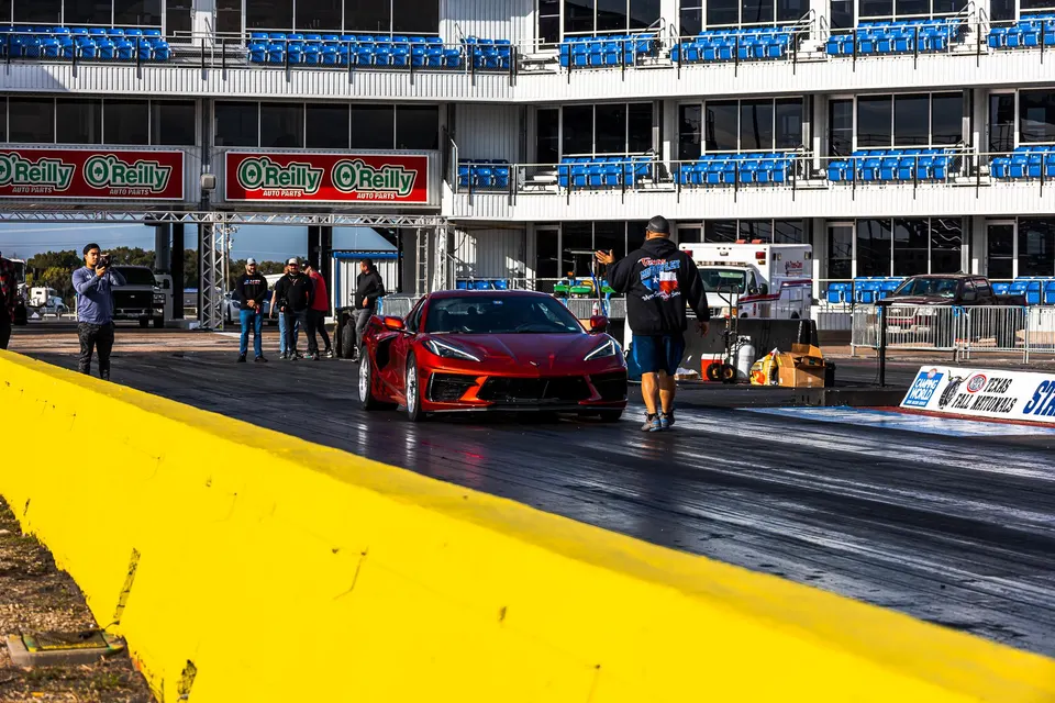 A red C8 Corvette at the starting line of a drag strip, with a crew member guiding it and spectators in the background.