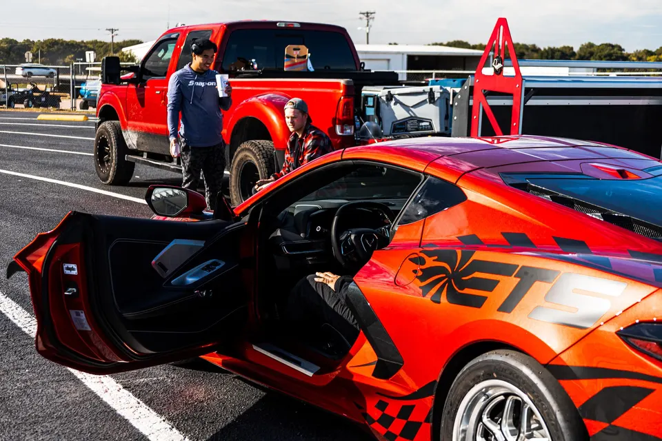 Red Chevrolet Corvette C8 with custom ETS livery at a drag racing event, parked next to a red Ford pickup truck with people.