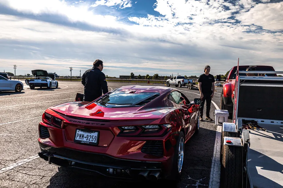 Red Corvette C8 in the staging lanes at a drag strip, with people standing around and other cars in the background.