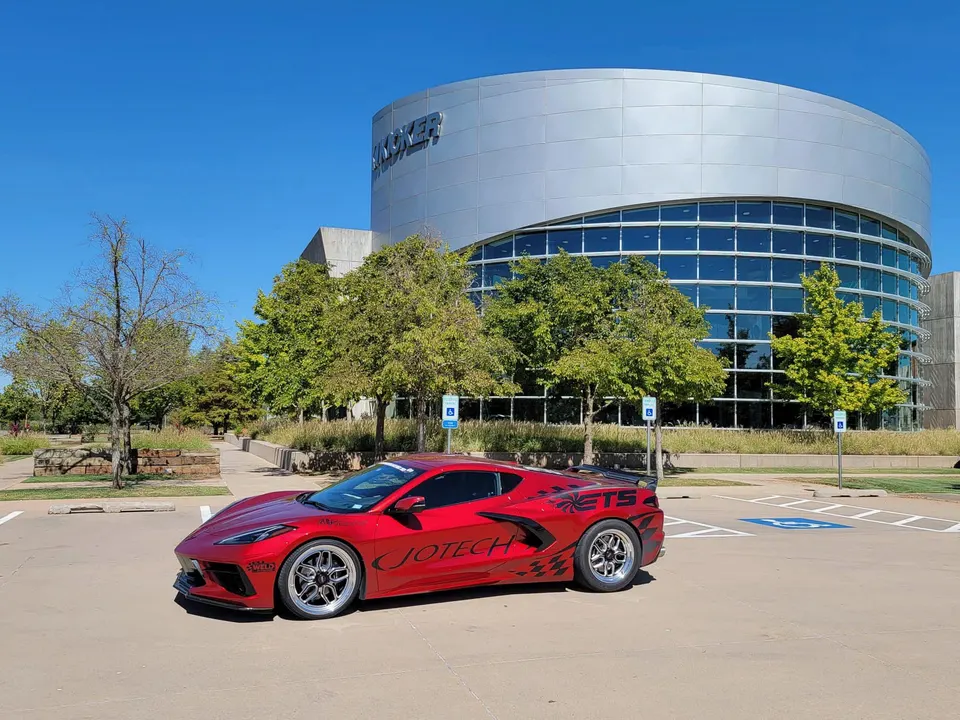 Red Corvette C8 with Jotech Motorsports livery parked in front of the Kicker building on a sunny day.