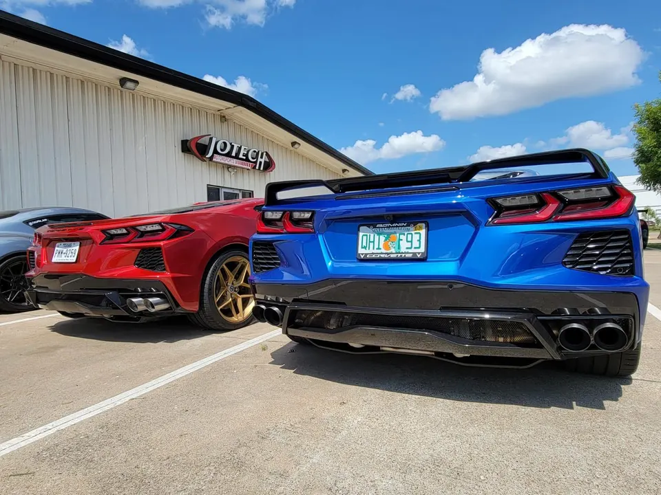 Two modified Chevrolet C8 Corvettes, one blue and one red, parked outside Jotech Motorsports with aftermarket rear wings and exhaust.