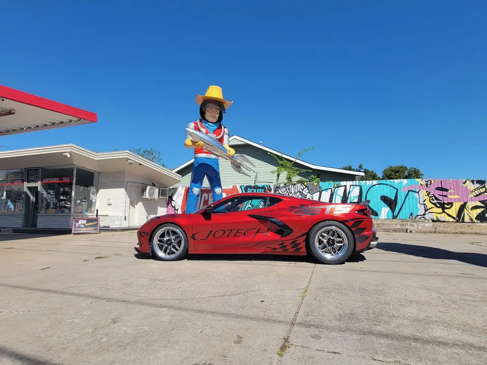 Red C8 Corvette with Jotech custom graphics and drag wheels parked next to a large statue and graffiti wall.
