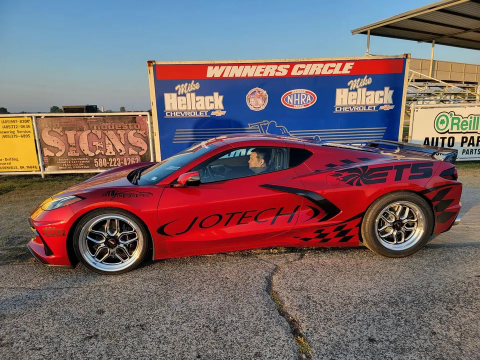 A red C8 Corvette drag car with custom wheels and Jotech livery parked at a drag strip next to a 'Winners Circle' banner.