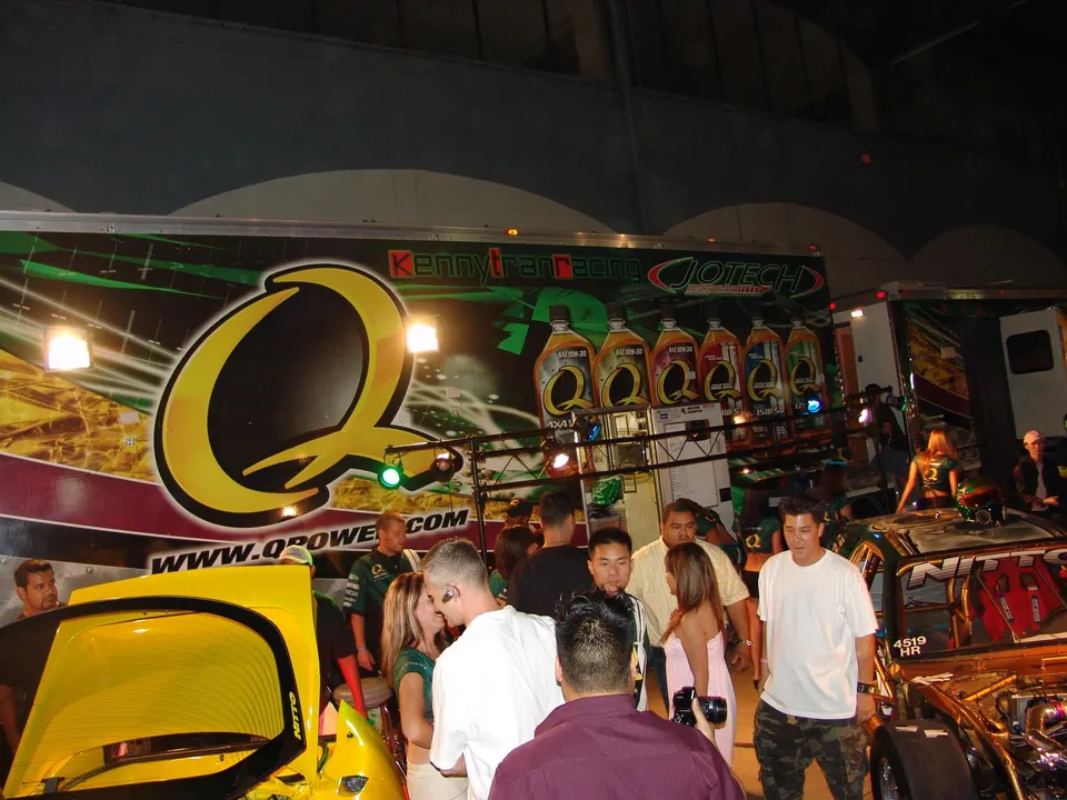 A crowd of people gathered around display cars at an event, with a large Kenny Tran Racing and Q Power banner in the background.