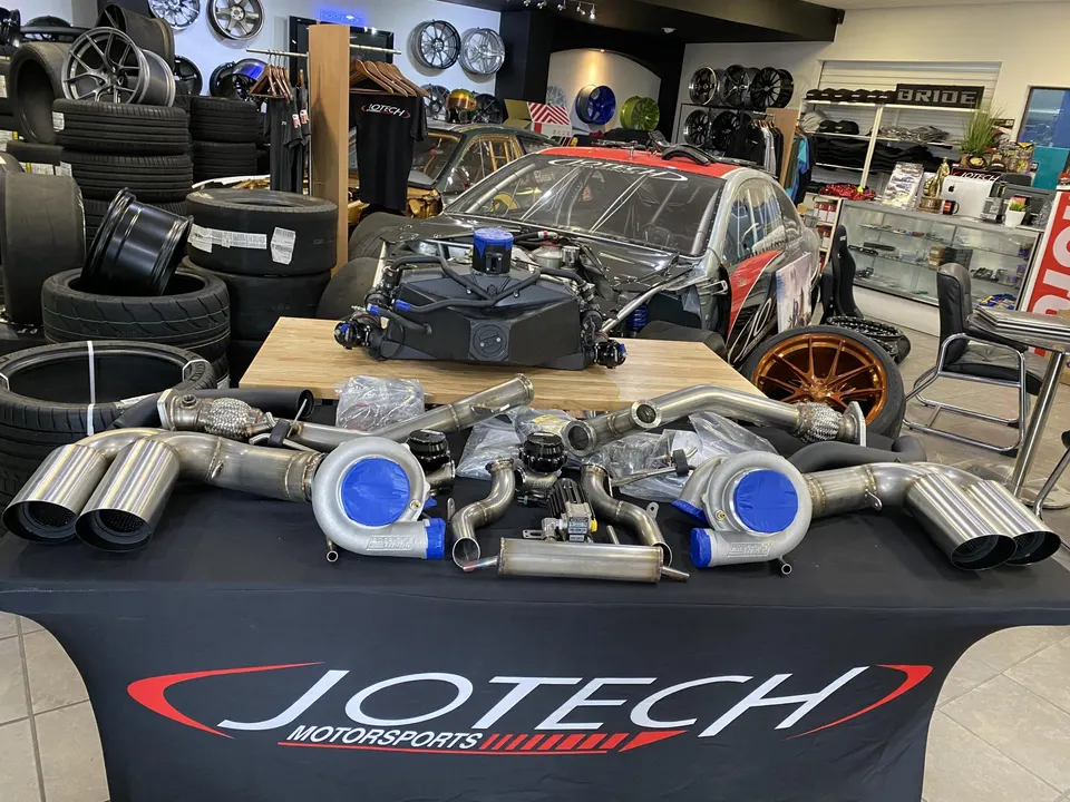 Jotech Motorsports shop with a twin-turbo kit, exhaust system, and engine on display, alongside a race car and stacked tires.