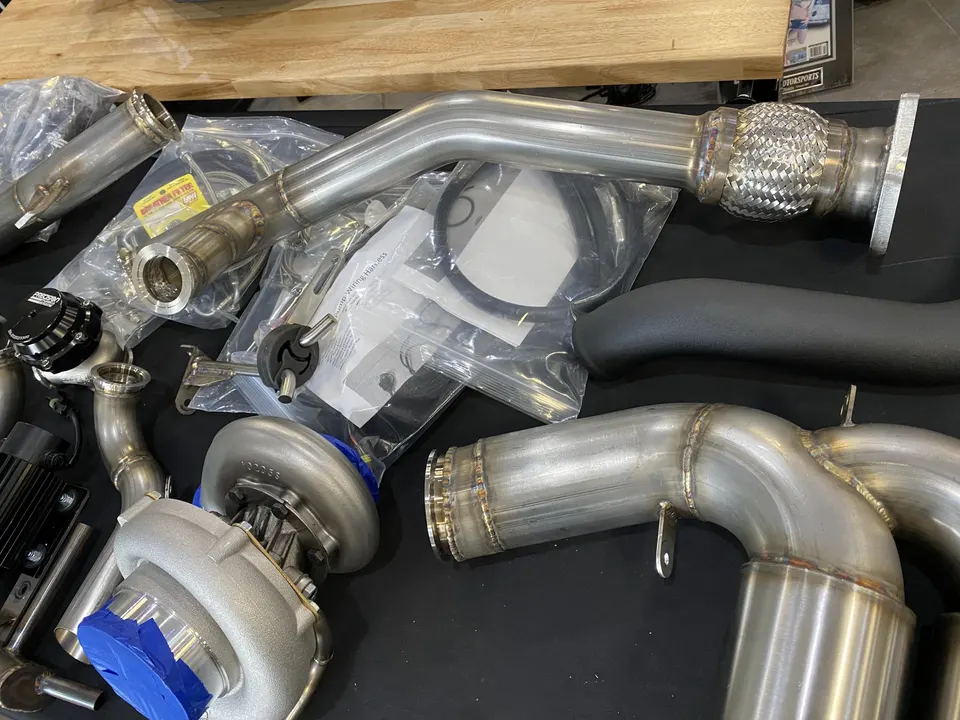 Assortment of new performance parts including a turbocharger, wastegate, and stainless steel exhaust components on a shop bench.