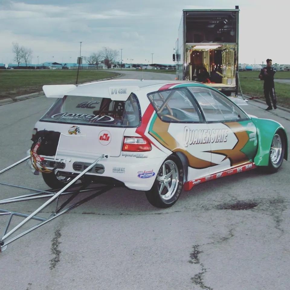 A white, green, and gold Honda Civic drag car with wheelie bars at a track, next to a race trailer with its door open.