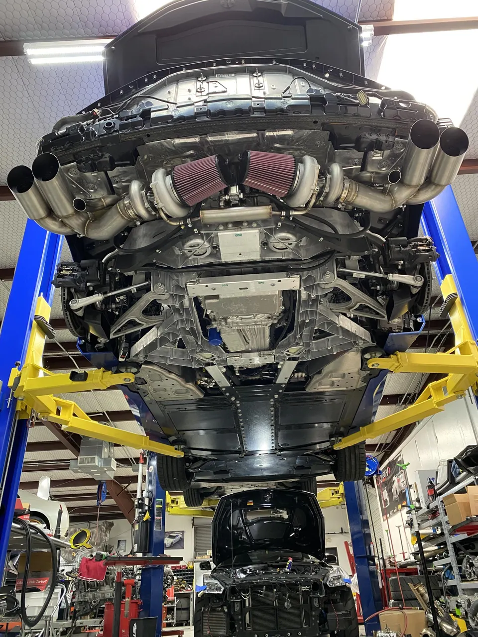Underside view of a C8 Corvette on a lift in a shop, showcasing a twin-turbo setup with large air filters and custom exhaust.