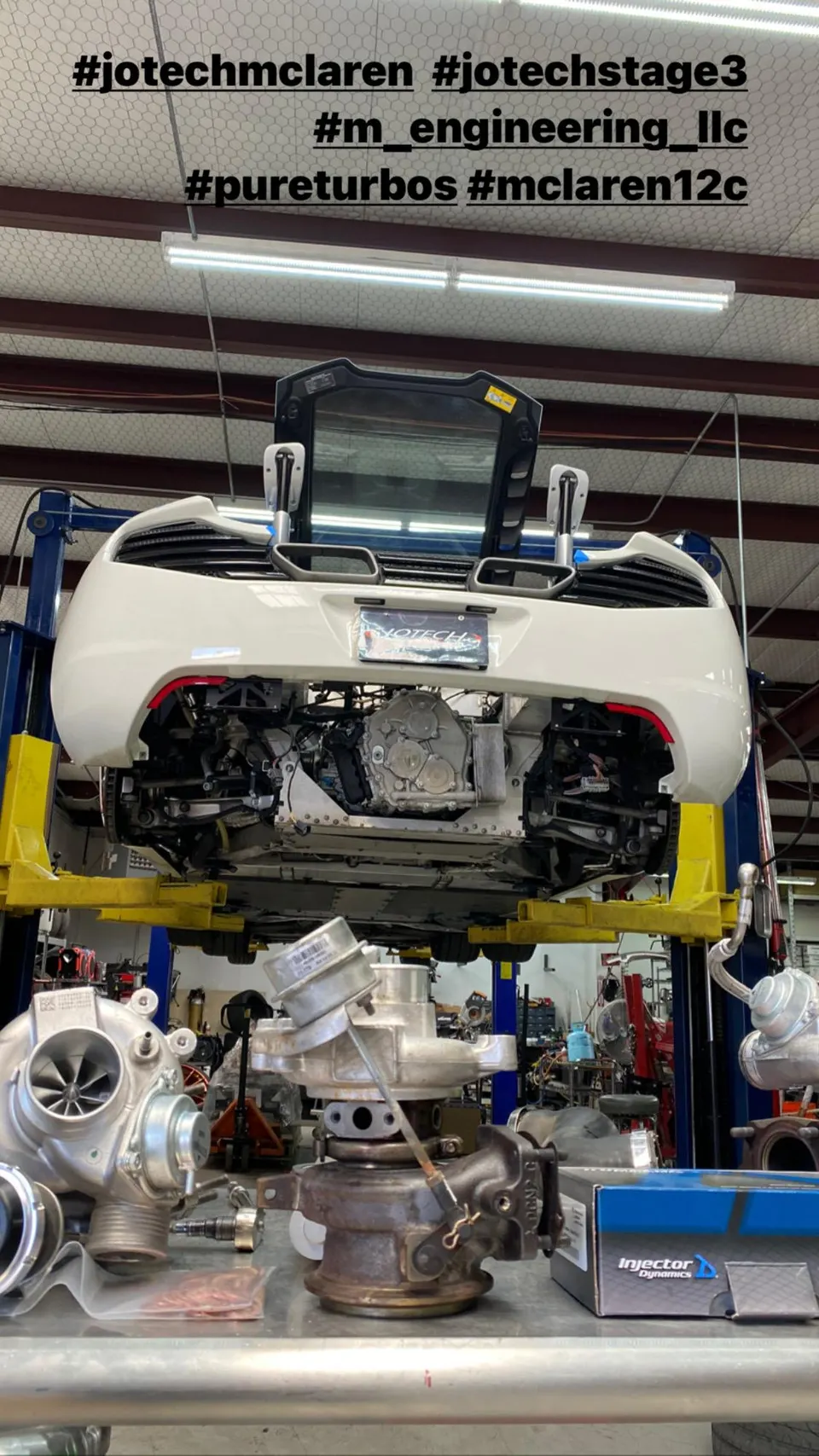 McLaren 12C on a lift in a performance shop with new and old turbochargers and fuel injectors on a workbench.
