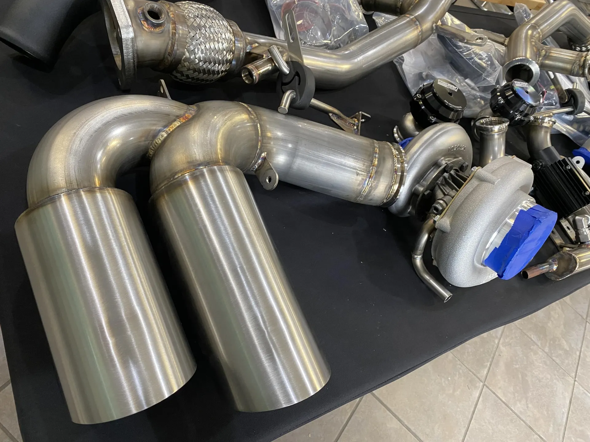 Collection of new stainless steel performance exhaust system components, turbocharger, and wastegates laid out in a shop.