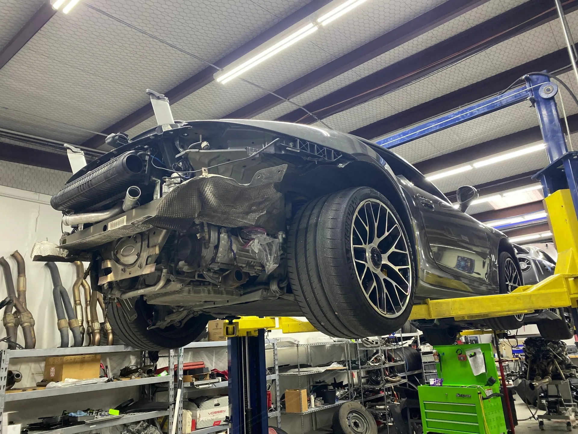 Porsche 911 on a lift in a performance shop with the rear bumper removed, exposing the exhaust system and engine components.