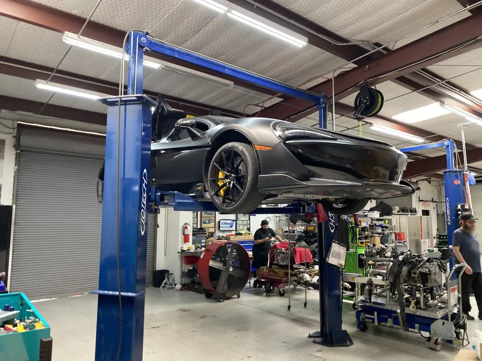 A gray McLaren supercar on a blue car lift in a Jotech Motorsports performance tuning shop with mechanics working.