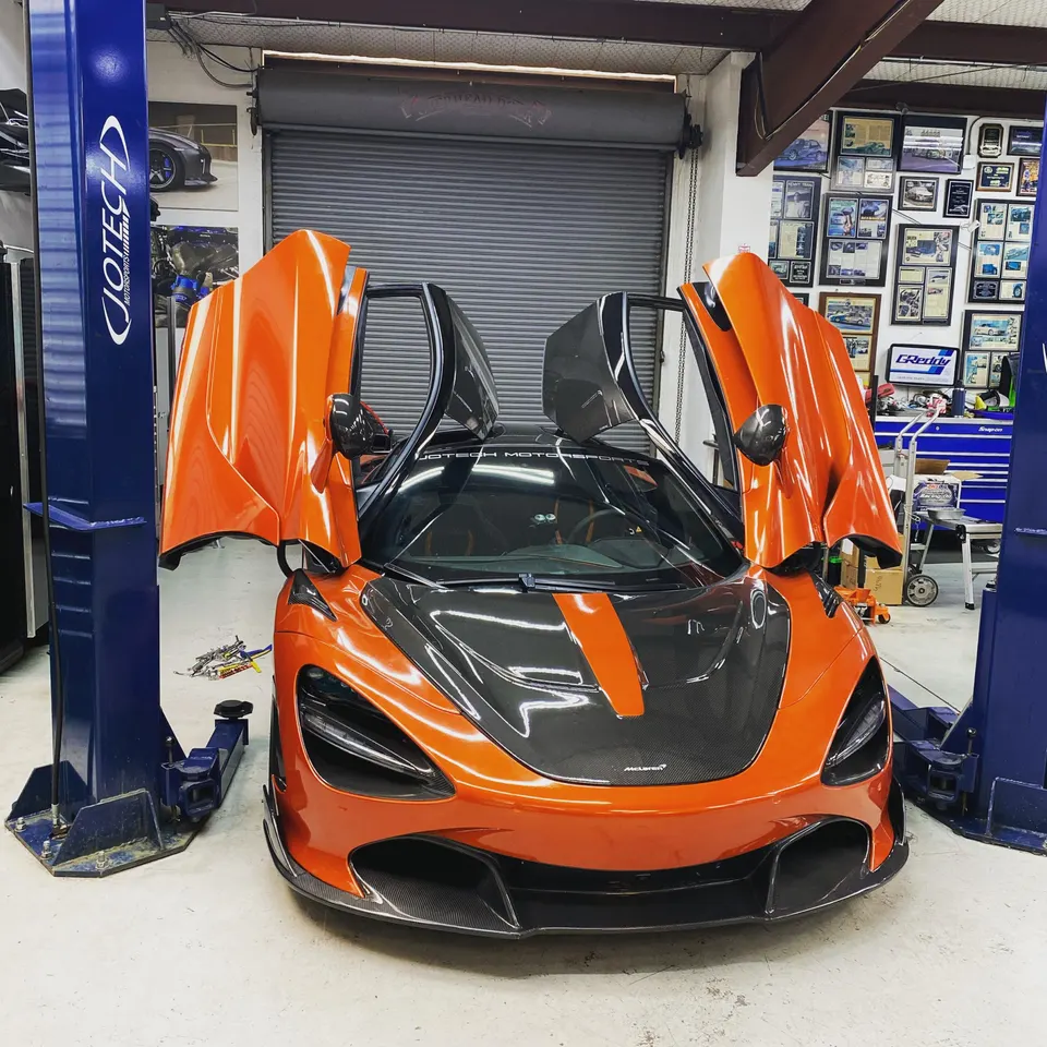 An orange McLaren 720S with its scissor doors open, parked in a Jotech Motorsports performance tuning shop.