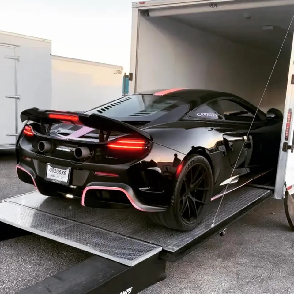 Black McLaren 570S with a large rear wing and pink accents being unloaded from a white enclosed trailer.