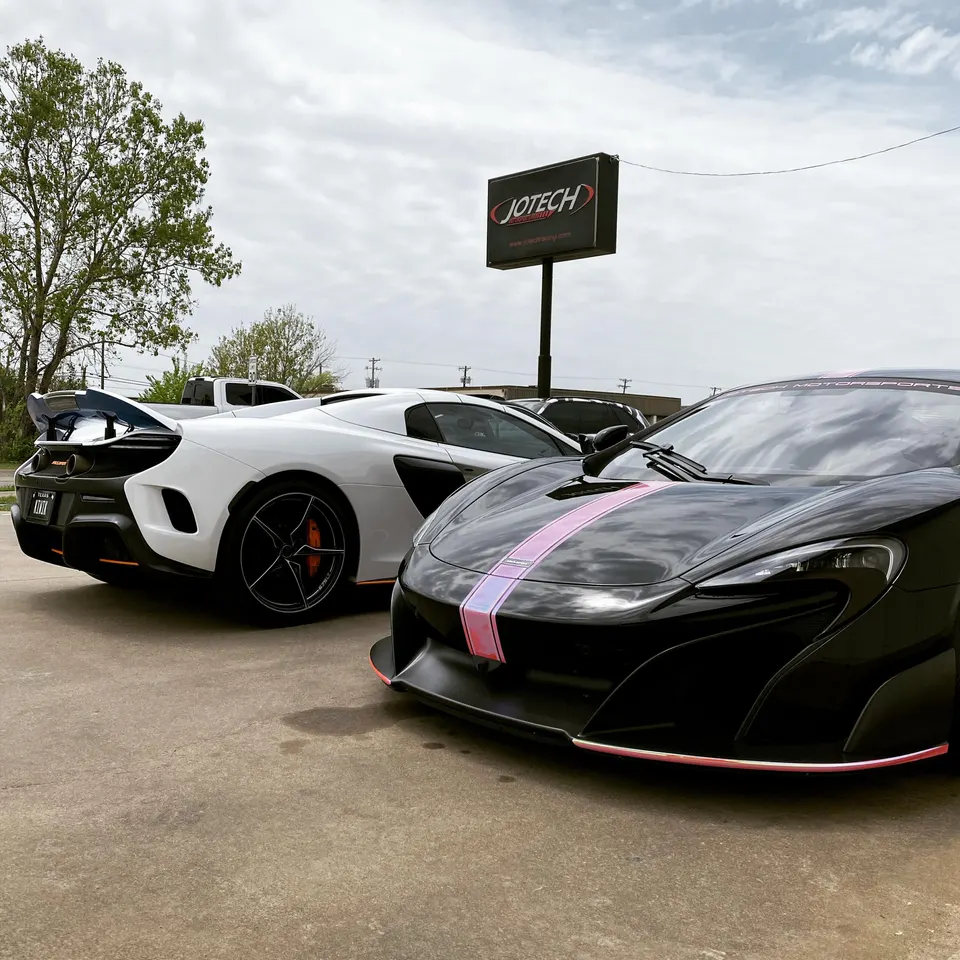 Two McLarens, one black with pink stripes and one white, parked outside Jotech Motorsports performance tuning shop.