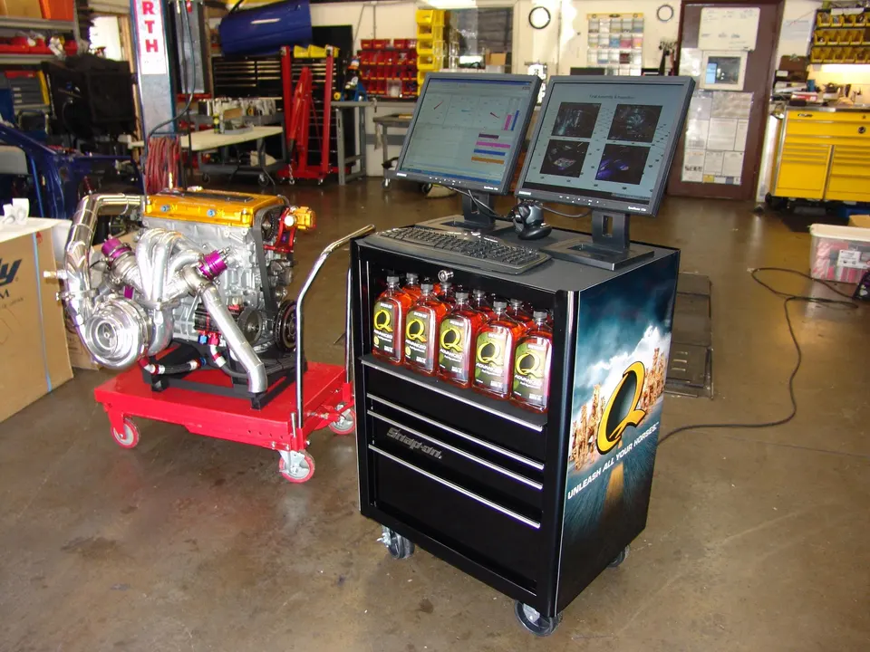 Highly modified performance engine with large turbocharger system on a cart in a tuning shop, next to a computer station.