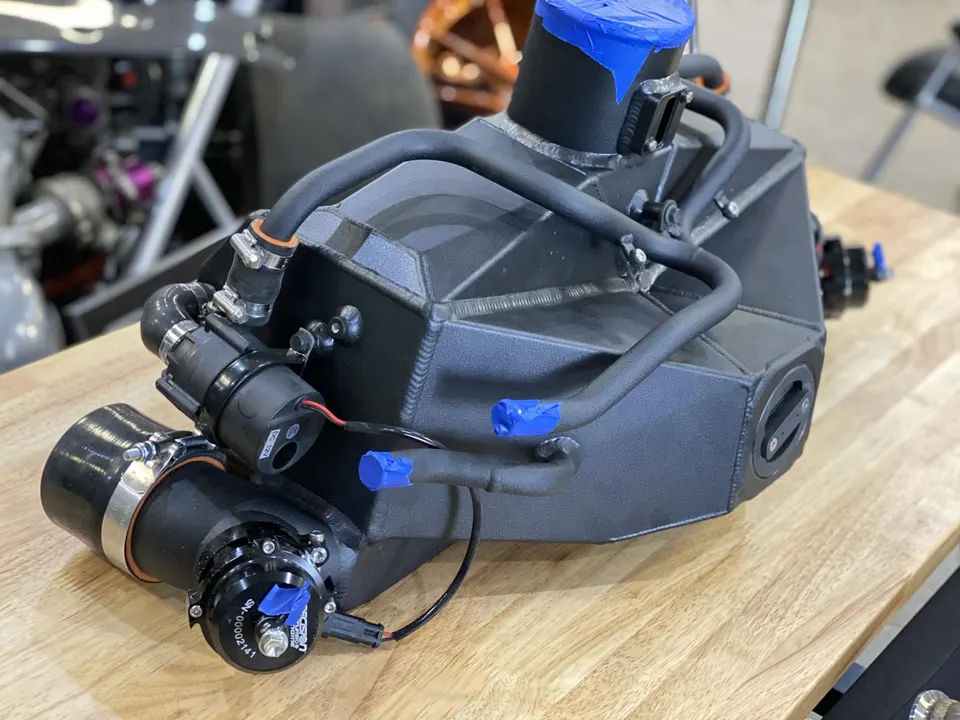 Close-up of a custom fabricated intercooler and coolant tank assembly with boost piping and blow-off valve on a workbench.