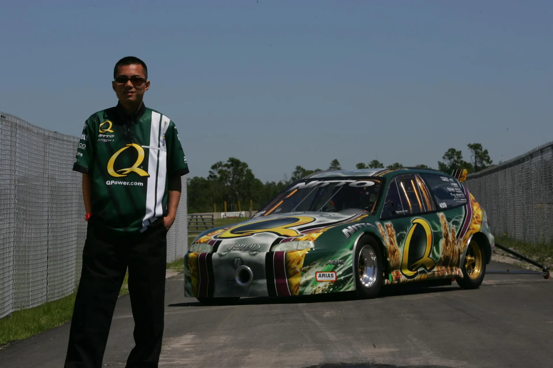 A person in a green Q Power shirt stands next to a green, gold, and white Q Power drag racing Honda Civic at a track.
