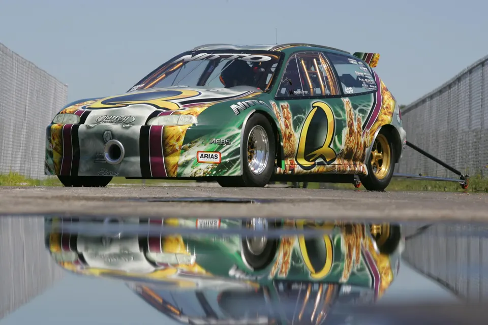 A custom-liveried Honda Civic drag car with wheelie bars on a track, reflected in a puddle under a clear sky.