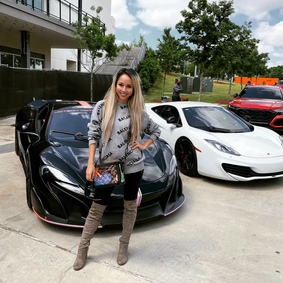 A woman posing with a black McLaren, a white McLaren, and a red Audi at an outdoor car event.