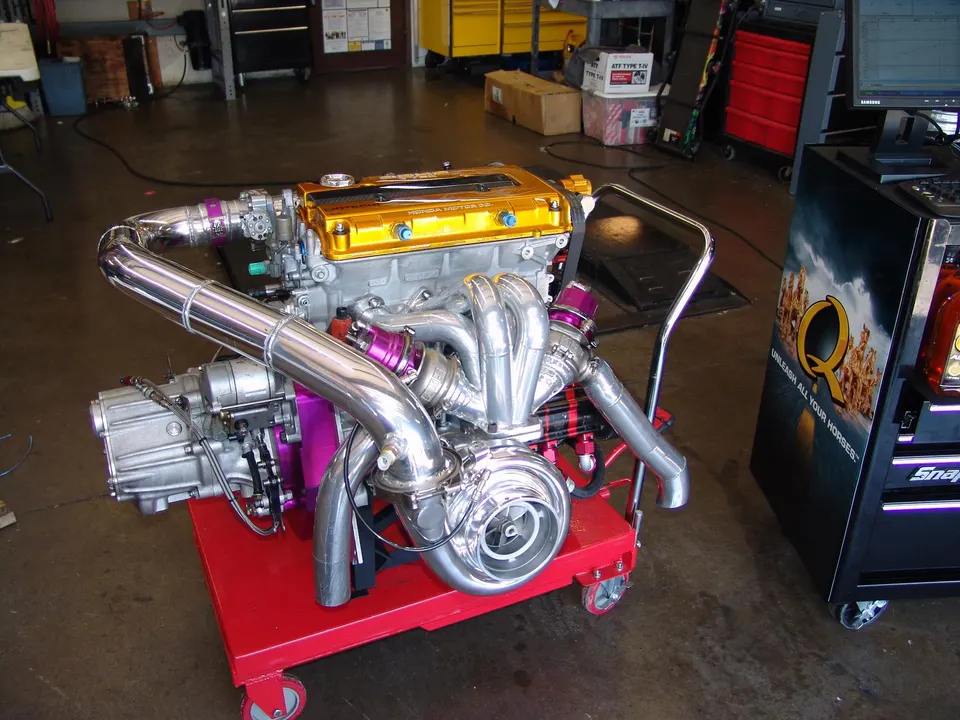 A highly modified Honda engine with a large turbocharger and custom exhaust manifold on an engine stand in a performance shop.