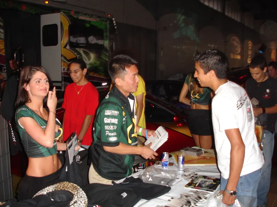 People interacting at a Jotech Motorsports booth during a car show event, with merchandise and a car in the background.
