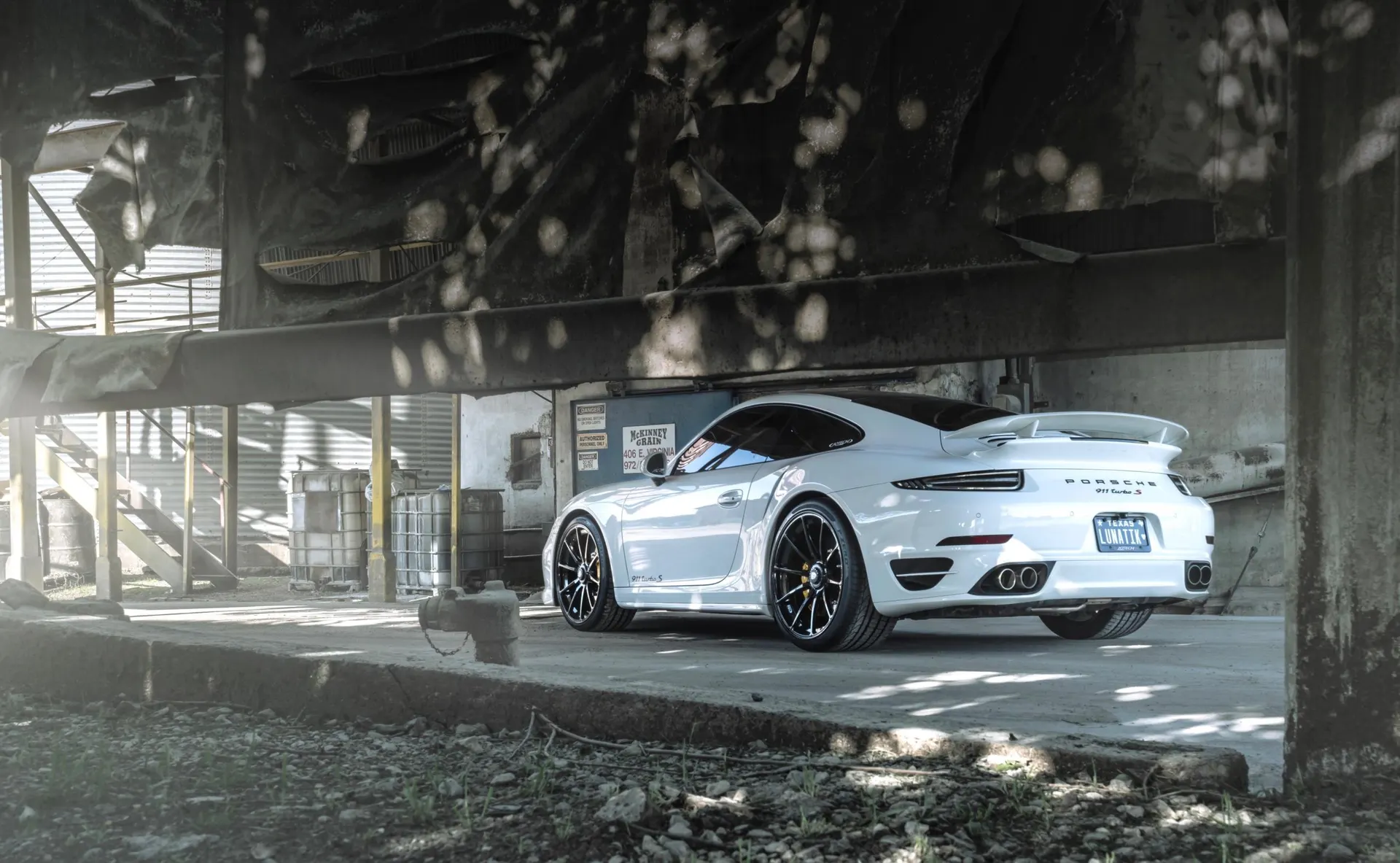 A white Porsche 911 Turbo S with aftermarket wheels parked in an industrial setting with dramatic lighting.