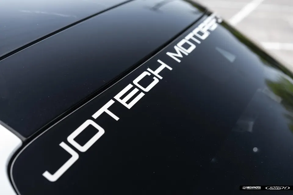 Close-up of a black car's rear window featuring a white Jotech Motors decal.