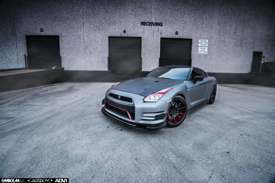 A matte grey Nissan GT-R with a carbon fiber hood and aftermarket wheels parked in front of a warehouse loading dock.