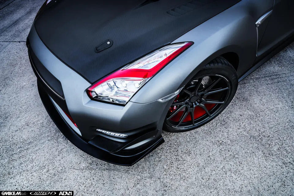 Matte grey Nissan GT-R with carbon fiber hood, aggressive front splitter, black aftermarket wheels, and red brake calipers.